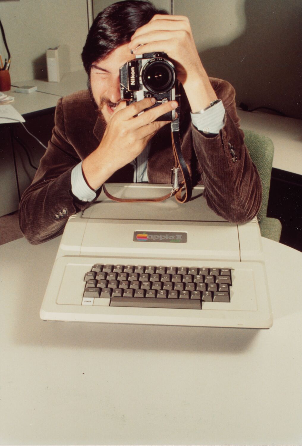 Steve peers through the lens of a Nikon camera as his elbow rests on the Apple II in front of him.
