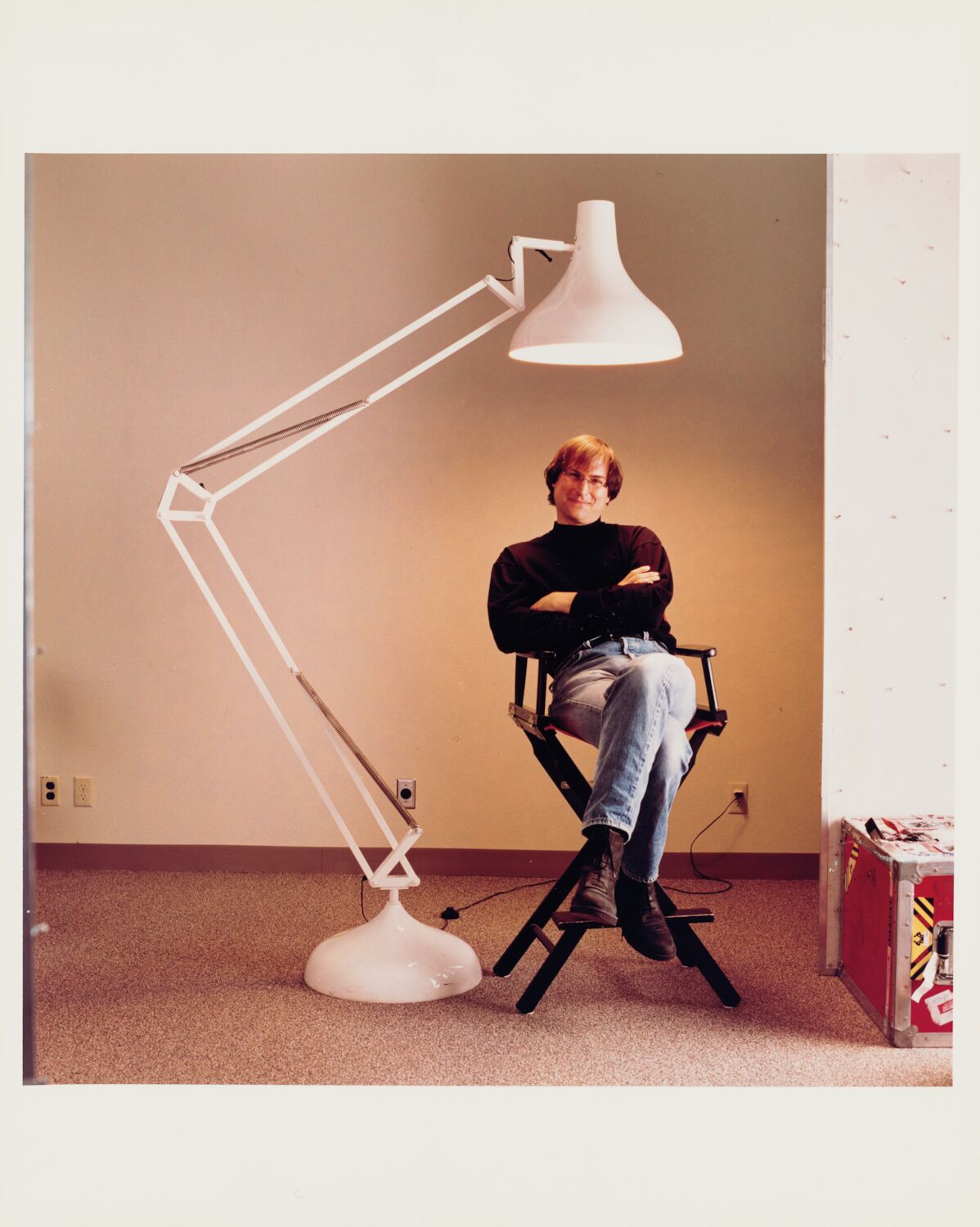 Steve, now wearing glasses, sits cross-legged in a director's chair under an oversized anglepoise Pixar-style lamp.