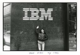 Steve, wearing a black leather jacket, holds his middle finger up toward a large IBM sign.