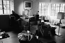 Steve sits cross-legged on the phone in his home office. The floor is strewn with boxes, papers, a tripod, and a trumpet.