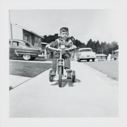 Two-year-old Steve rides a tricycle with tassels on the handlebars toward the camera.