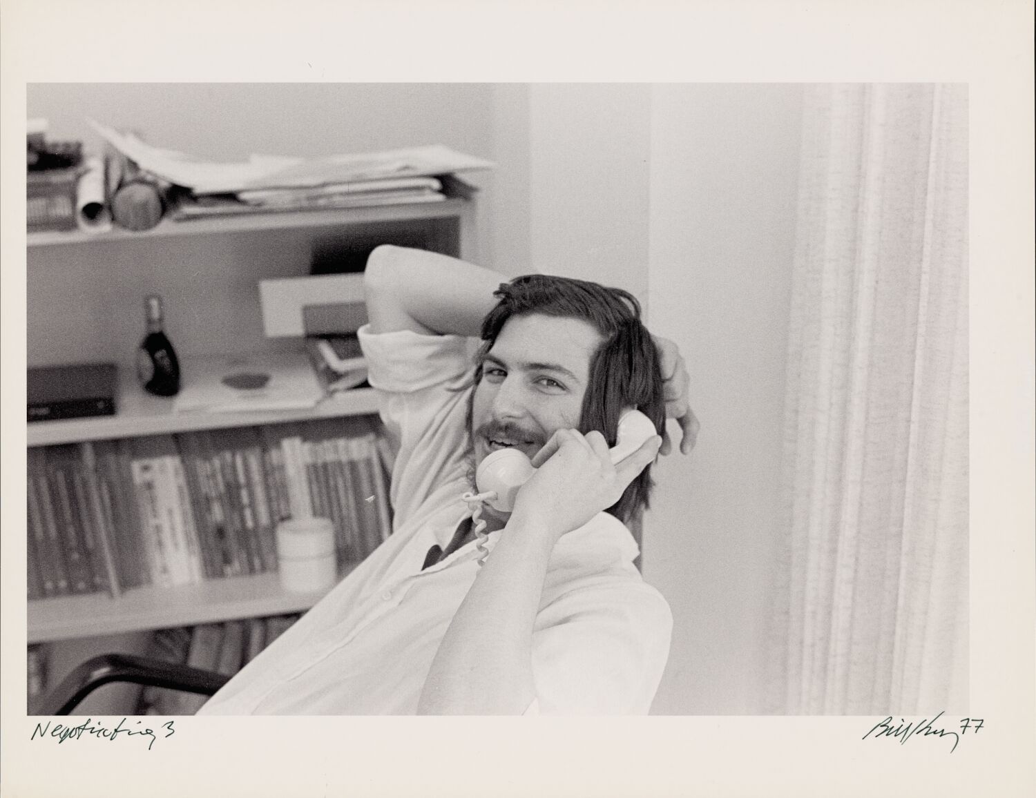 A scruffy-bearded Steve smiles at the camera as he leans back in his office chair and talks on the telephone.