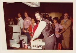 Wearing suit pants and a vest, Steve is in front of a small crowd, looking over his shoulder at the camera.