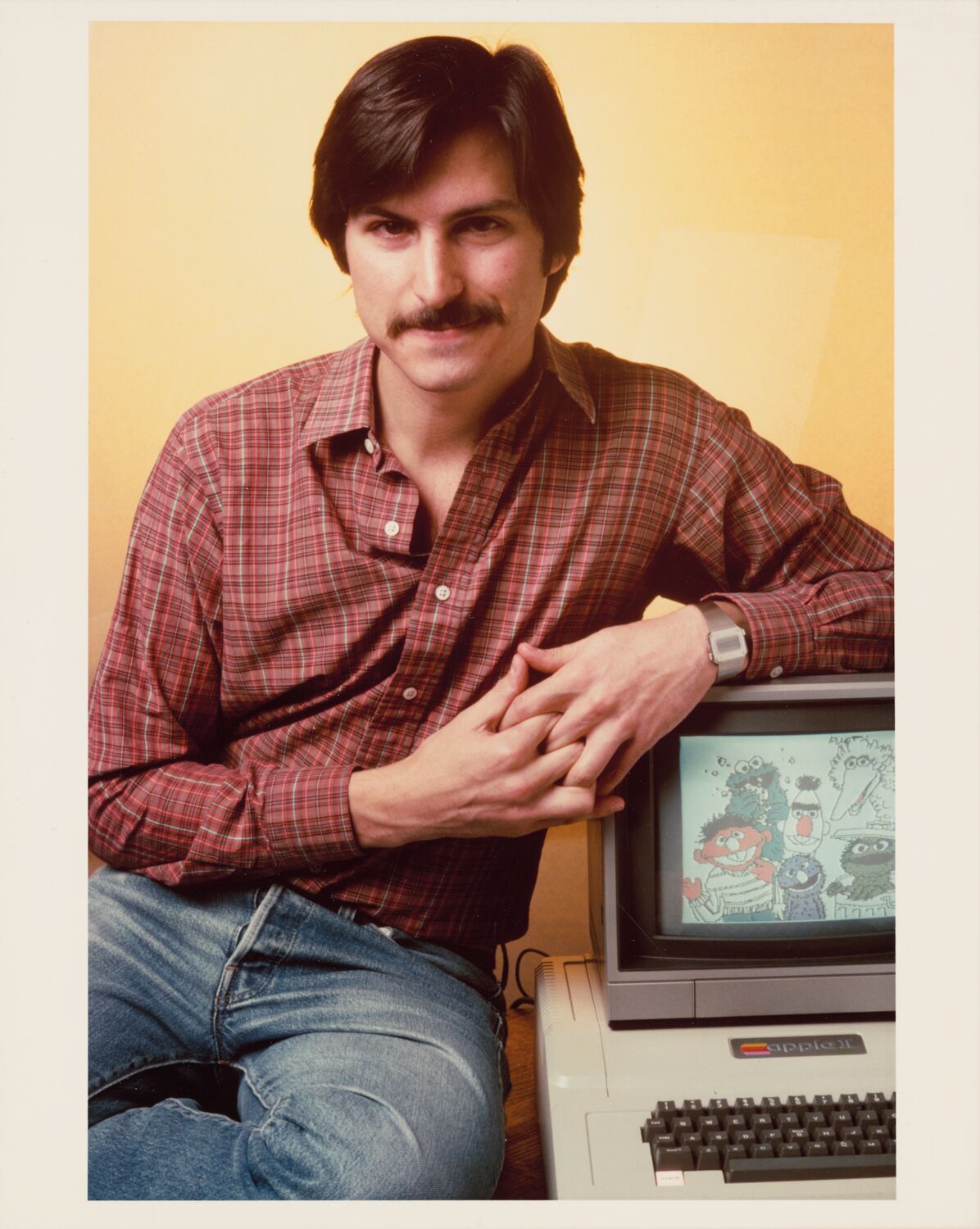 With tidy hair and a plaid shirt, Steve poses with an Apple II displaying drawings of Sesame Street characters.
