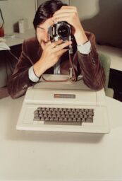 Steve peers through the lens of a Nikon camera as his elbow rests on the Apple II in front of him.