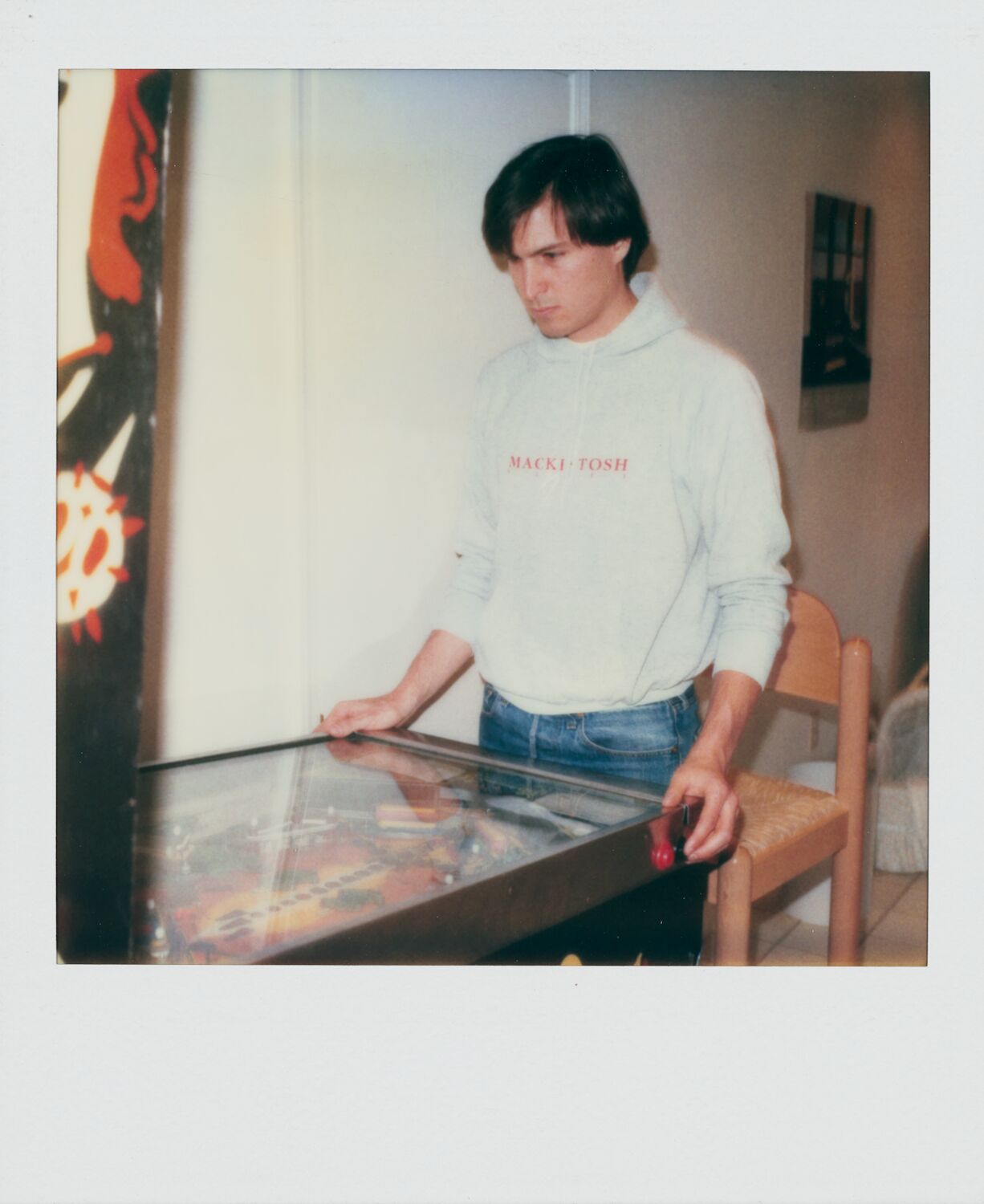 A Polaroid of Steve, playing pinball, with a white hooded sweatshirt with the word Mackintosh on the chest.