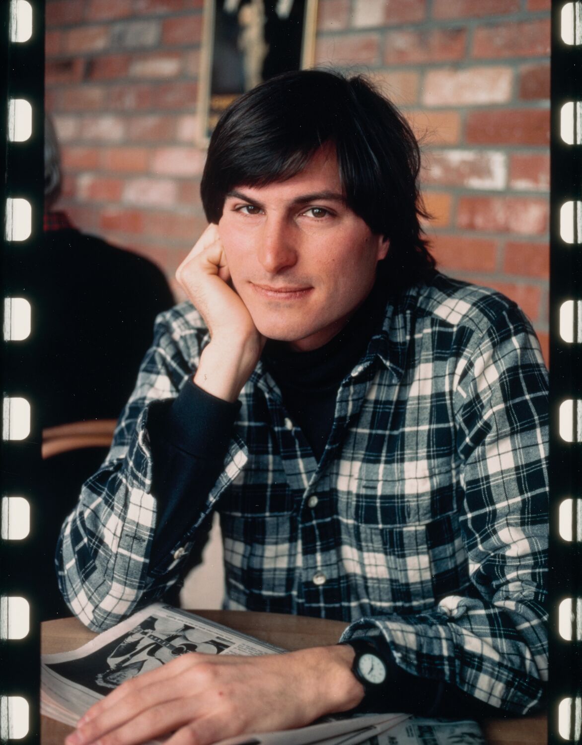 Steve gazes into the camera as he cradles his chin in his right hand, his left hand resting on top of a newspaper on the table.