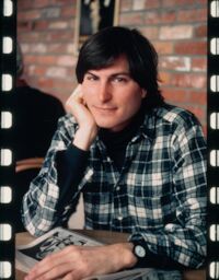 Steve gazes into the camera as he cradles his chin in his right hand, his left hand resting on top of a newspaper on the table.