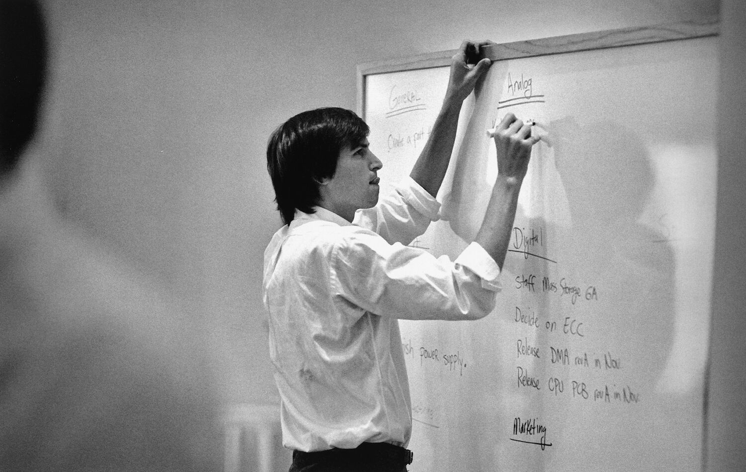 Steve, in a white shirt, writes on a whiteboard. He has a look of concentration and is sticking out his tongue.