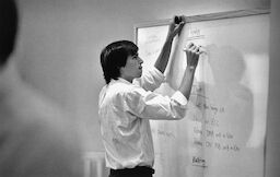 Steve, in a white shirt, writes on a whiteboard. He has a look of concentration and is sticking out his tongue.