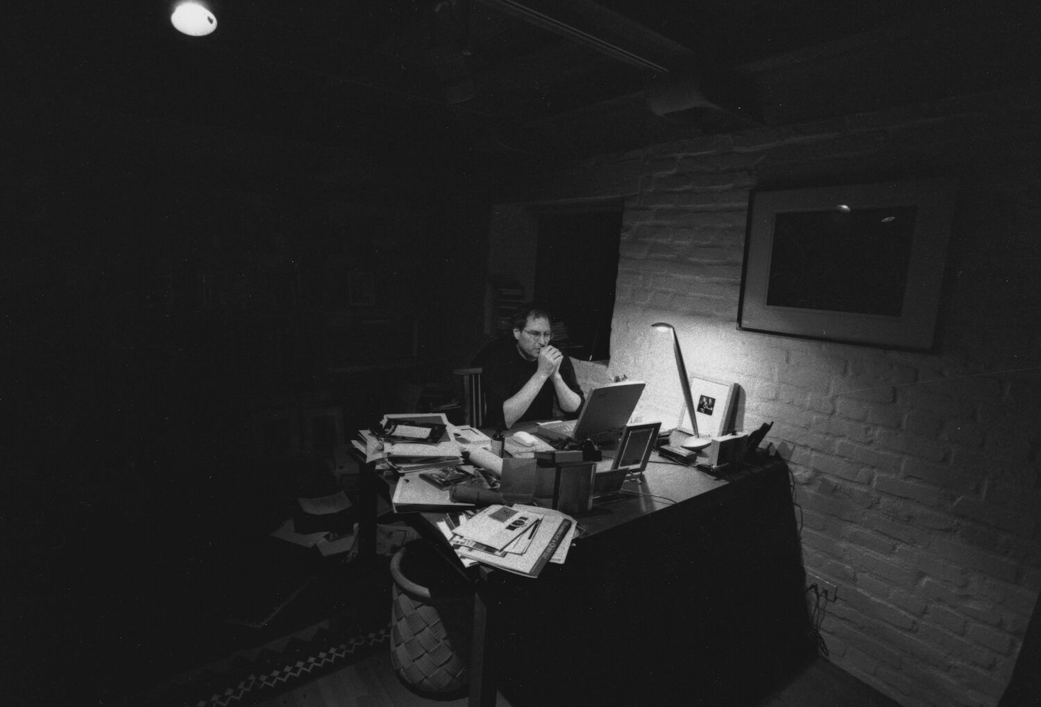 In his dark home office, a small lamp illuminates Steve as he sits at a cluttered desk.