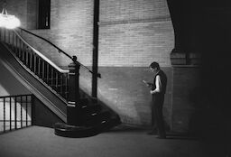 Steve stands in a dimly-lit stairwell opposite a large brick wall. He wears a vest over a white shirt.