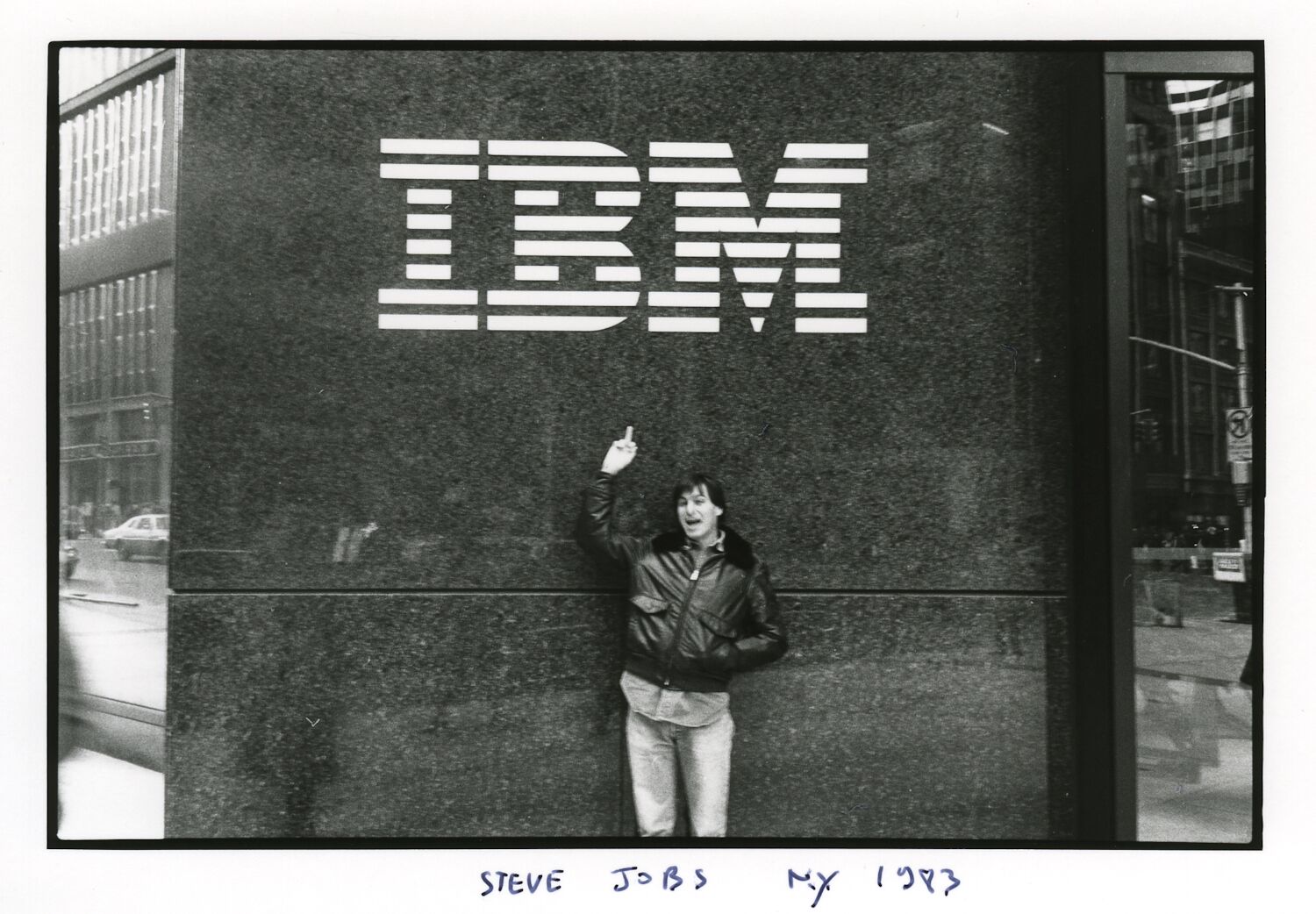 Steve, wearing a black leather jacket, holds his middle finger up toward a large IBM sign.