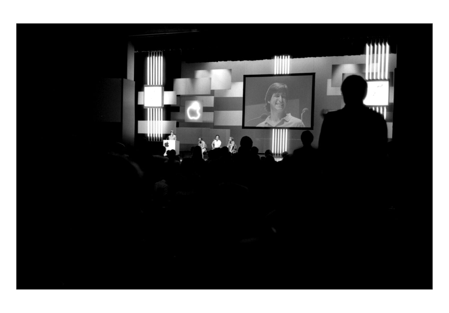 Viewed through a conference crowd, Steve - his clean-shaven, grinning face - is displayed on a large screen.