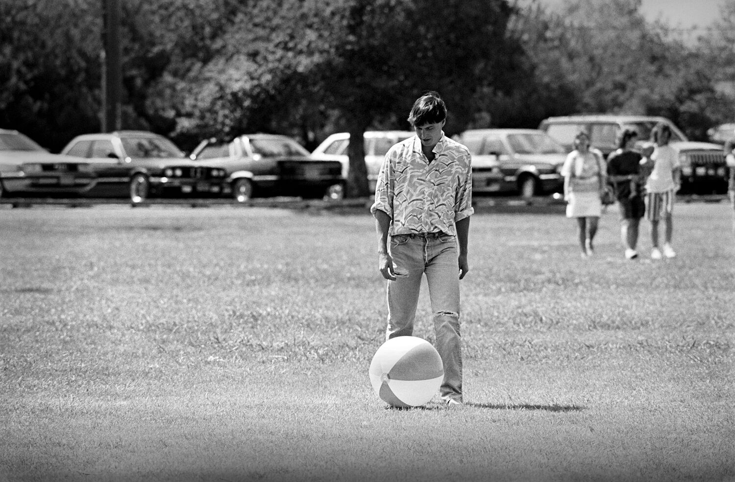 Steve kicks a beach ball in a grassy park. He is dressed in ripped jeans and a patterned shirt with rolled-up sleeves.