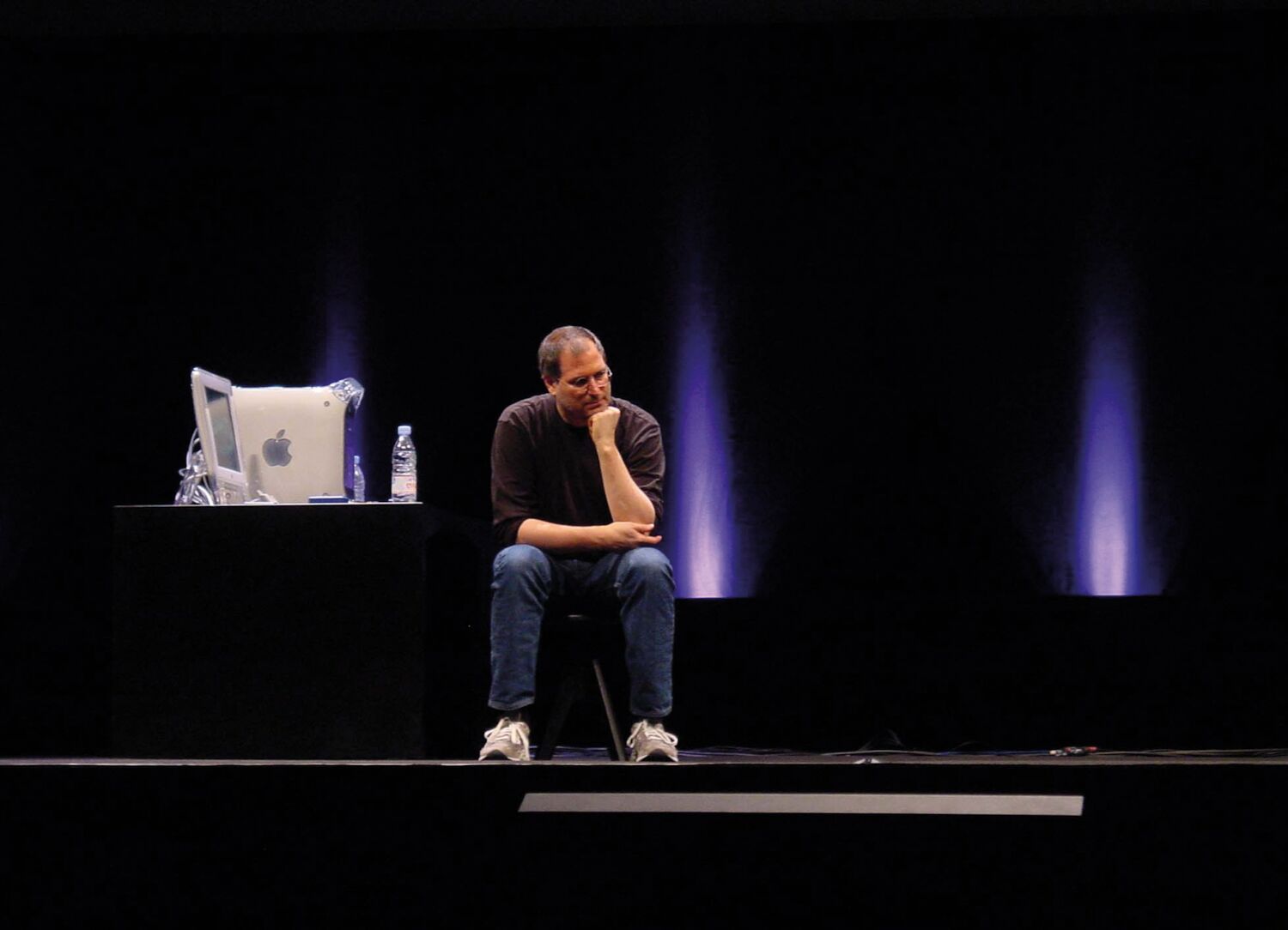 Steve takes a break during stage rehearsals for MacWorld. Sitting on a stool, he leans forward with his hand on his chin.
