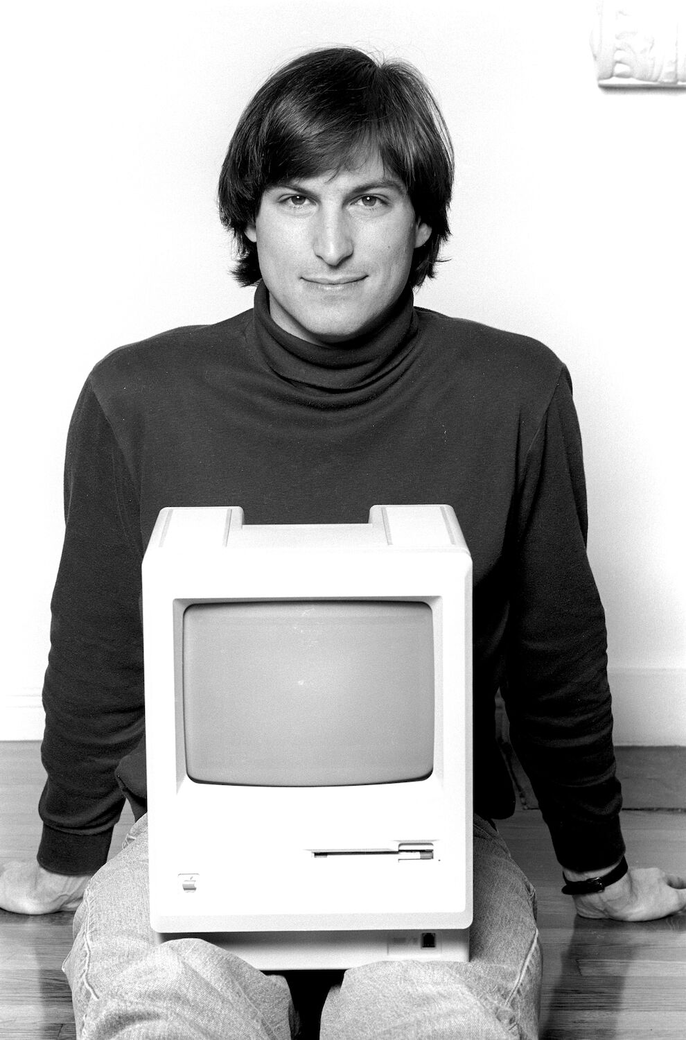 29-year-old Steve wearing a black turtleneck and jeans, poses with a Macintosh computer resting in his lap.