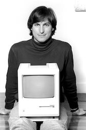 29-year-old Steve wearing a black turtleneck and jeans, poses with a Macintosh computer resting in his lap.