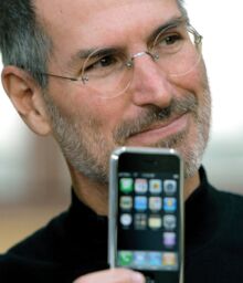 Steve holds a first generation iPhone up to his face and smiles; the phone is out of focus.