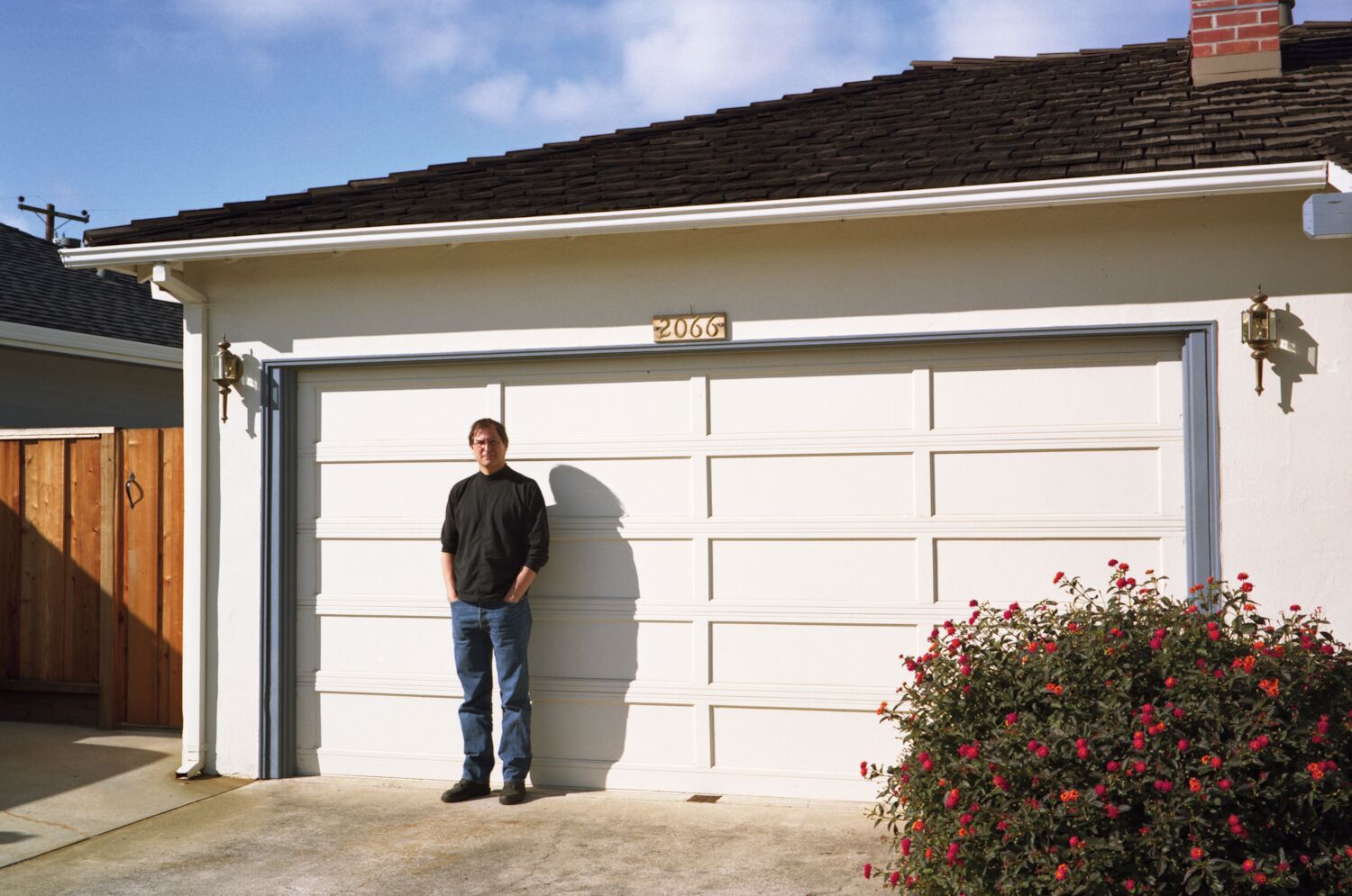 Steve stands against the white garage door of the house where Apple started in 1976. The house number reads 2066.