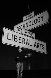 Steve is on stage. Behind him are two oversized, intersecting street signs: one says Technology, the other Liberal Arts.