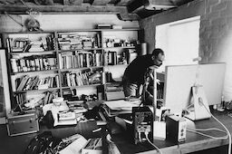 Steve leans across a cluttered desk to use his Mac. Behind him, shelves are piled with books and boxes litter the floor.