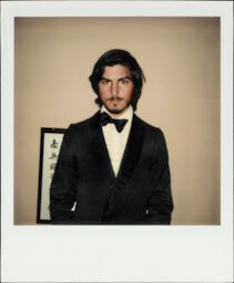 A Polaroid of young Steve, with long hair and mustache, wearing a tuxedo with a lop-sided bow tie.