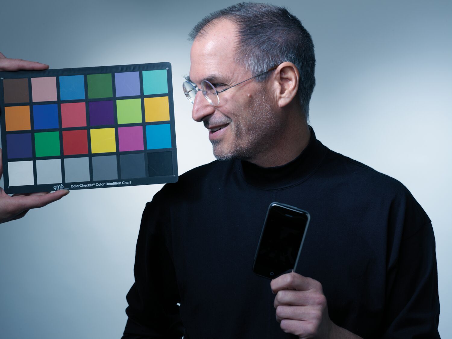 Steve, holding an iPhone, smiles and looks to the side as a pair of hands enters the frame, holding a color chart.