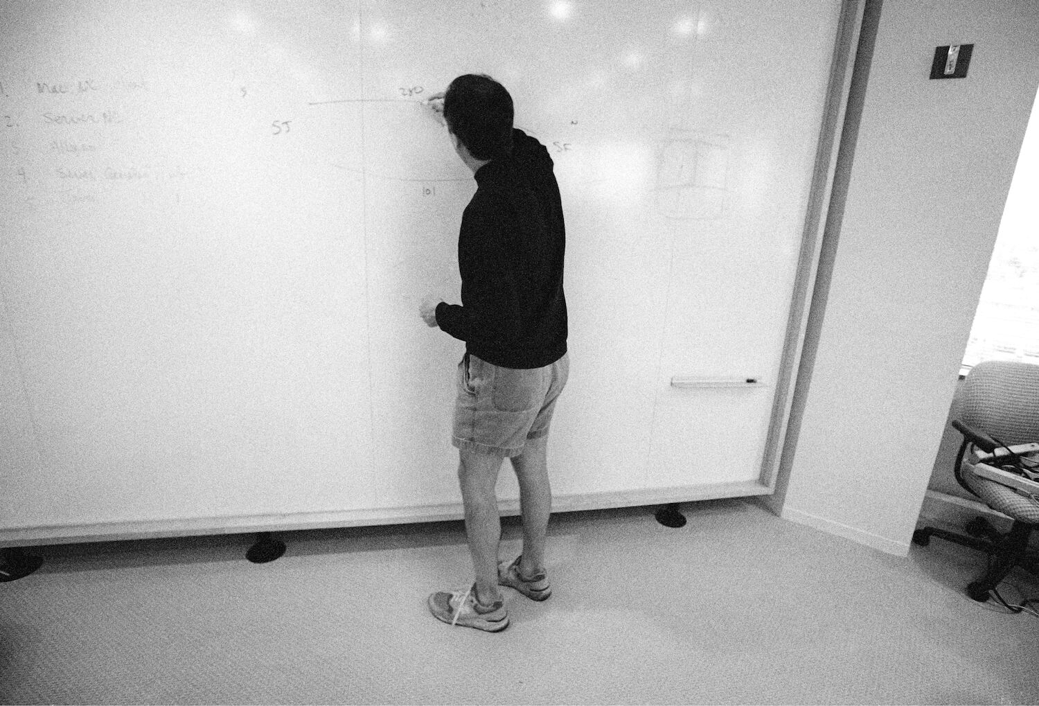 Steve has his back turned to the camera as he writes on a whiteboard. He wears a black turtleneck, shorts and sneakers.